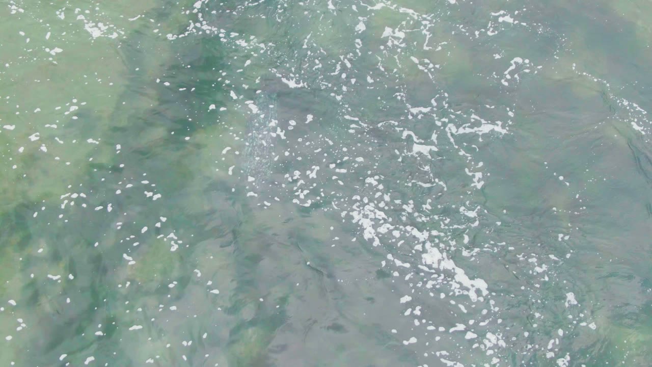 Pale green ocean water in La Bocana fills the frame, with broken foam flecks, submerged dark patches, and no visible people.