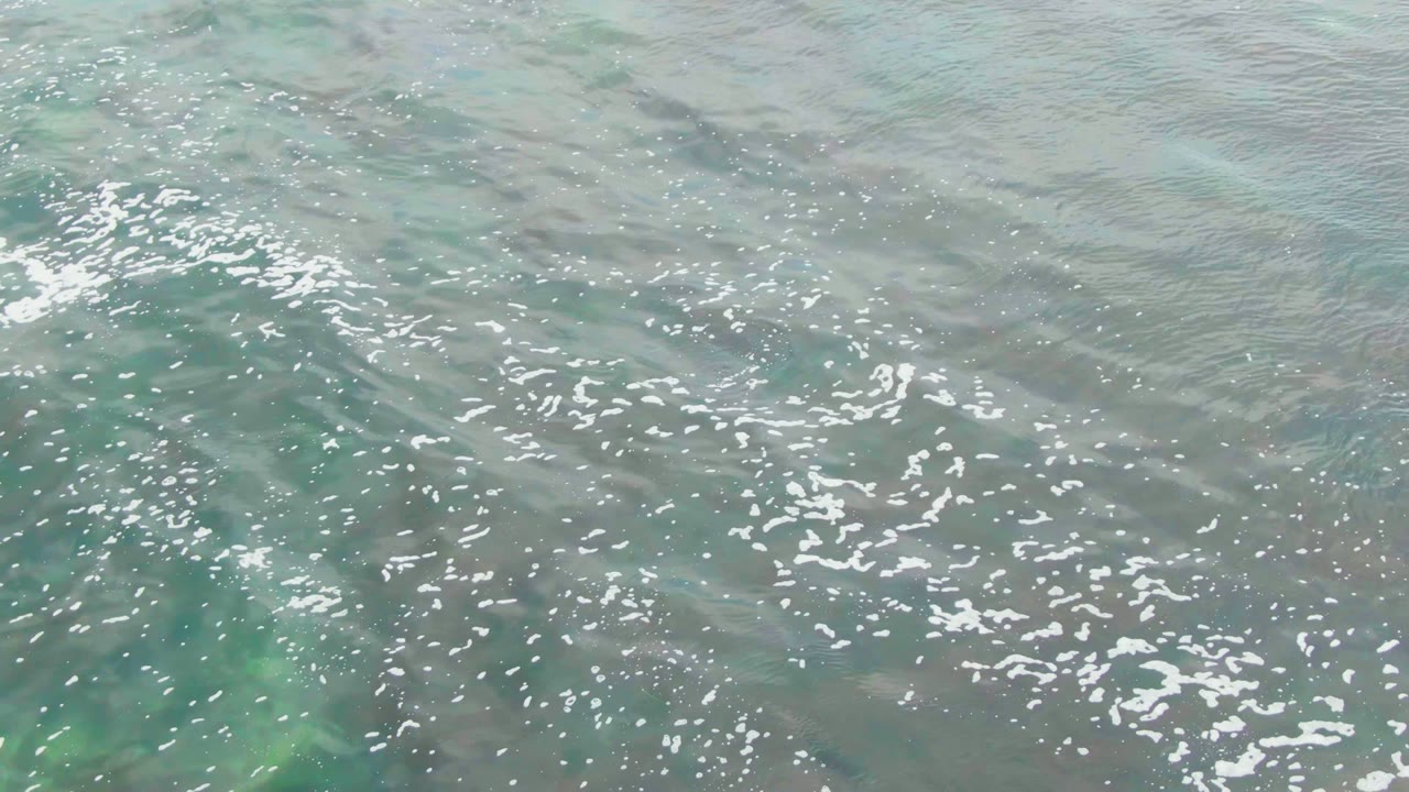 Clear shallow ocean water in La Bocana, Baja California Sur, with scattered white foam across pale turquoise and green surface tones, no visible people.