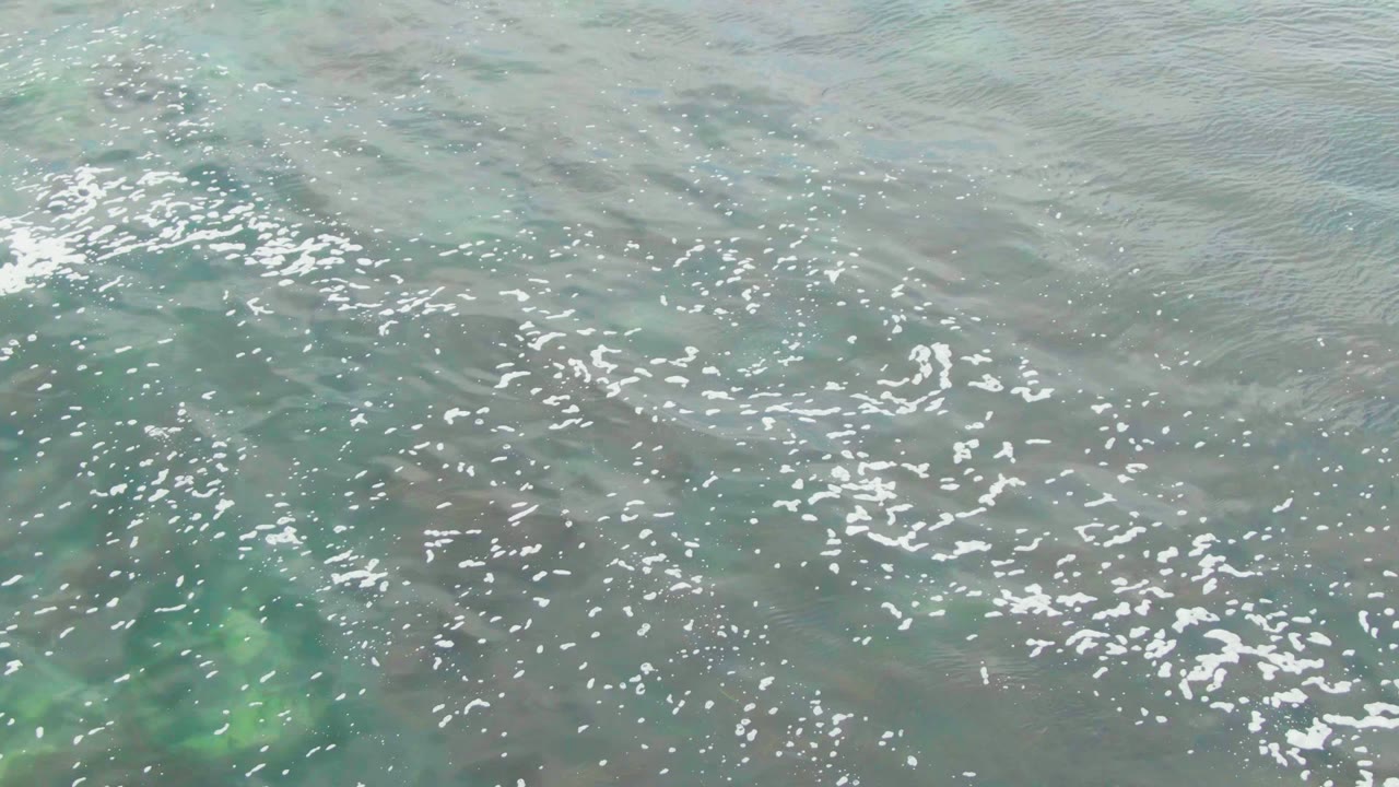 Clear ocean surface at La Bocana with dense white foam flecks near the upper center and soft green patches showing through the shallow water, no visible people.