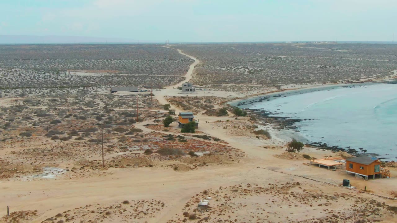 Aerial frame over La Bocana, Baja California Sur showing a sandy dirt road through scrub, a small orange structure, utility poles, blue bay water, rocky shoreline, and no visible people.