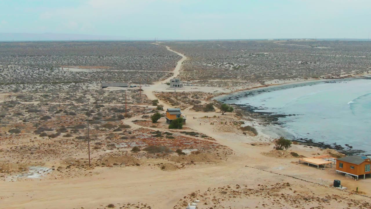Coastal desert road with small structures and pale blue water along a rocky shore.