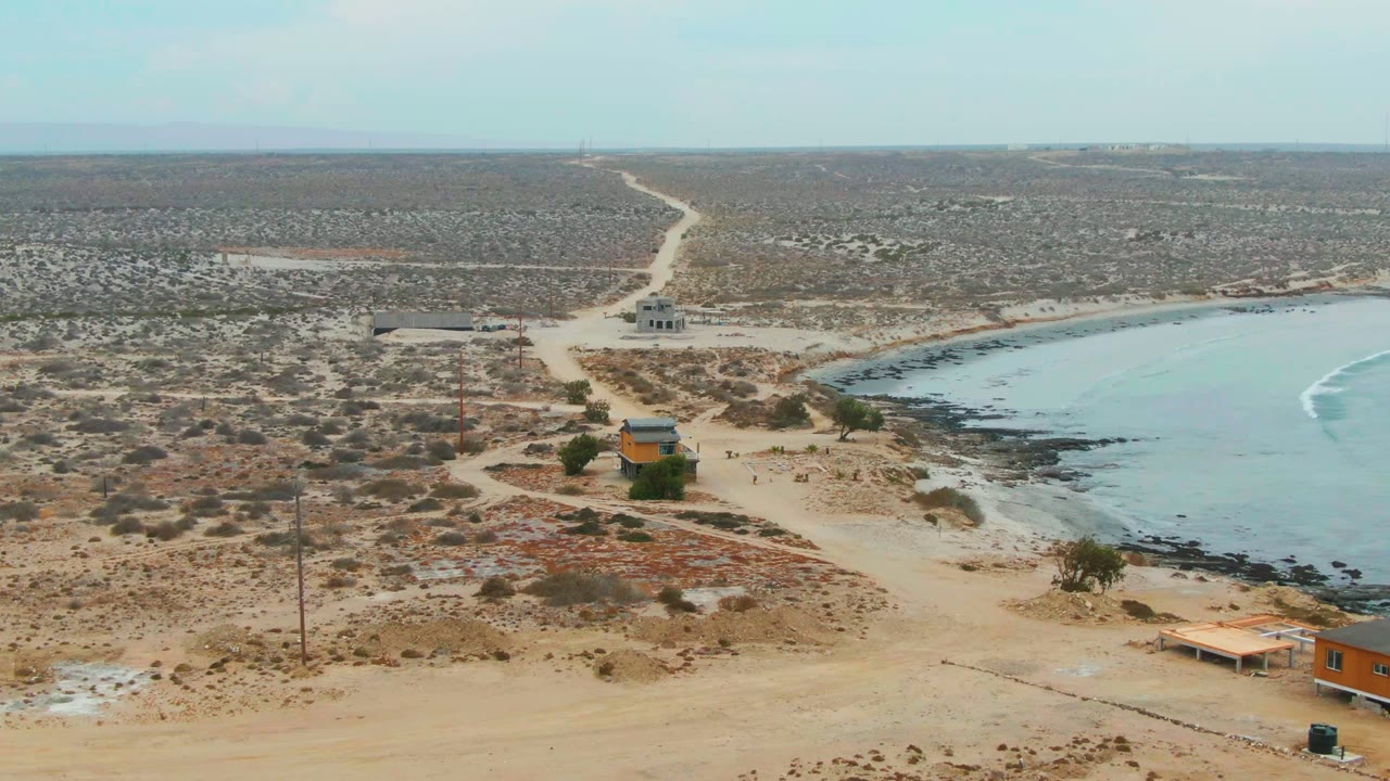 La Bocana aerial frame with sandy tracks crossing dry vegetation, small buildings near the coast, a pale blue bay at right, a white surf line, and no visible people.