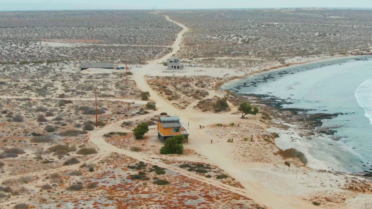 Opening aerial frame over La Bocana, Baja California Sur, showing a small orange building near sandy paths, scrub desert, rocky shoreline, pale blue water, and no visible people.