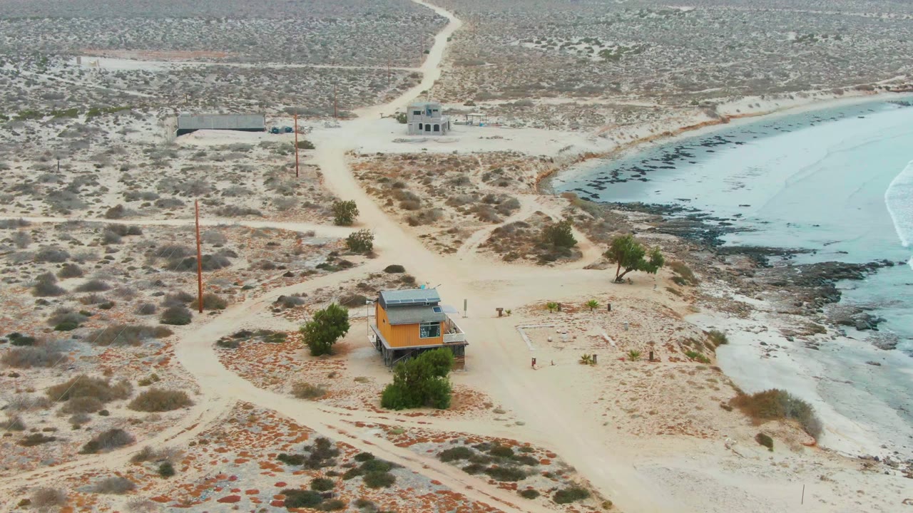 Mid-clip aerial view of La Bocana's desert edge, with sandy tracks curving around a small orange structure, blue water to the right, rocky shore texture, and no visible people.