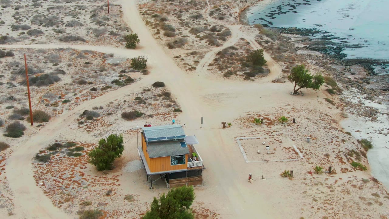 Aerial frame over La Bocana, Baja California Sur showing a small house with roof solar panels beside sandy roads, sparse vegetation, and rocky shoreline, with no visible people.