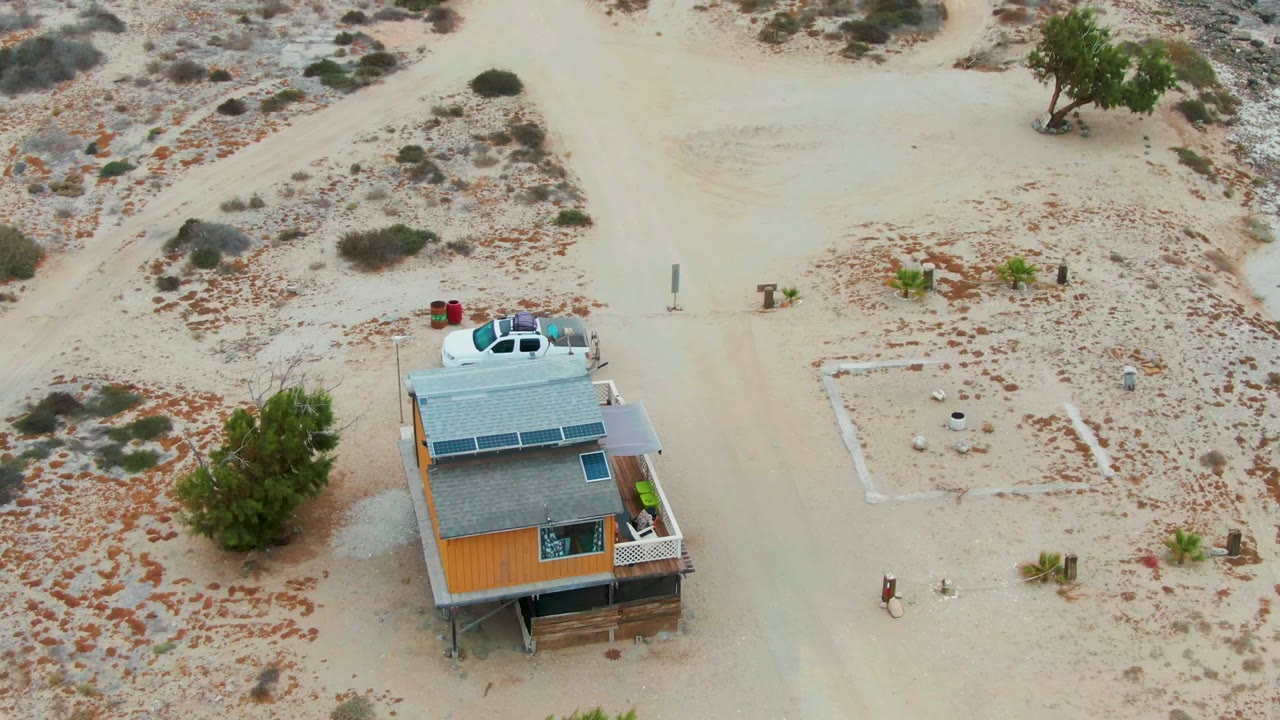 Drone frame over La Bocana, Baja California Sur showing the house and a white car near the roofline, with sandy roads, scrub vegetation, and no visible people.