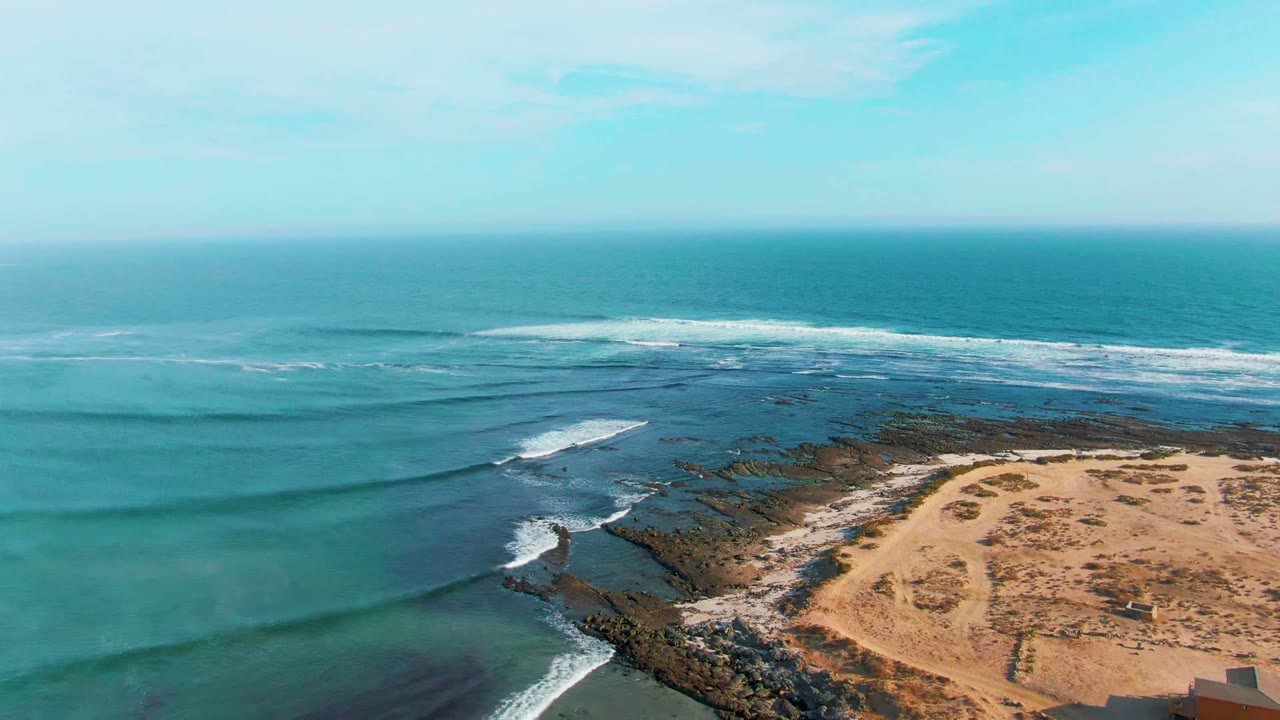 Aerial view over La Bocana, Baja California Sur, with turquoise water, white surf lines, dark rocks, sandy beach at lower right, and no visible people.