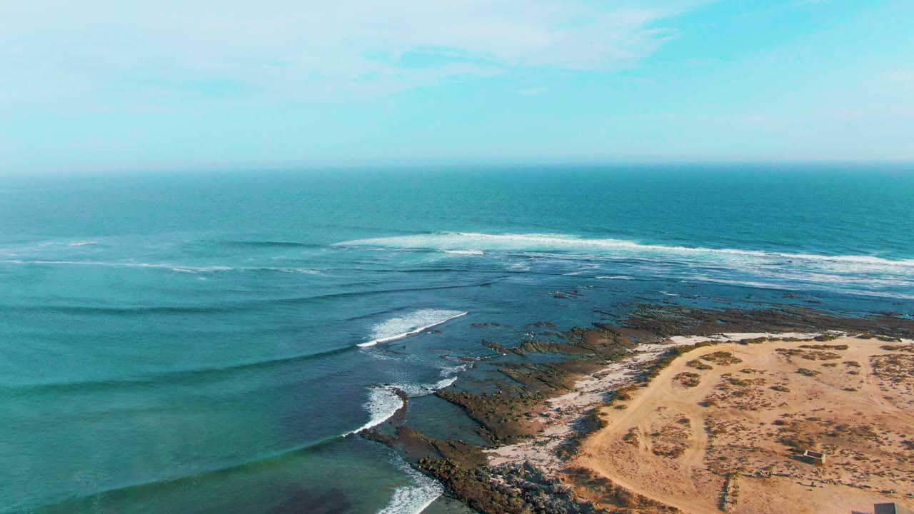 Aerial coastline view with waves curling toward dark rocks and sand.
