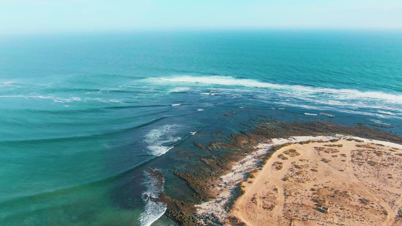 Aerial frame over La Bocana, Baja California Sur, showing turquoise water, white surf, dark rock shelves, pale sand, sparse scrub, and no visible people.
