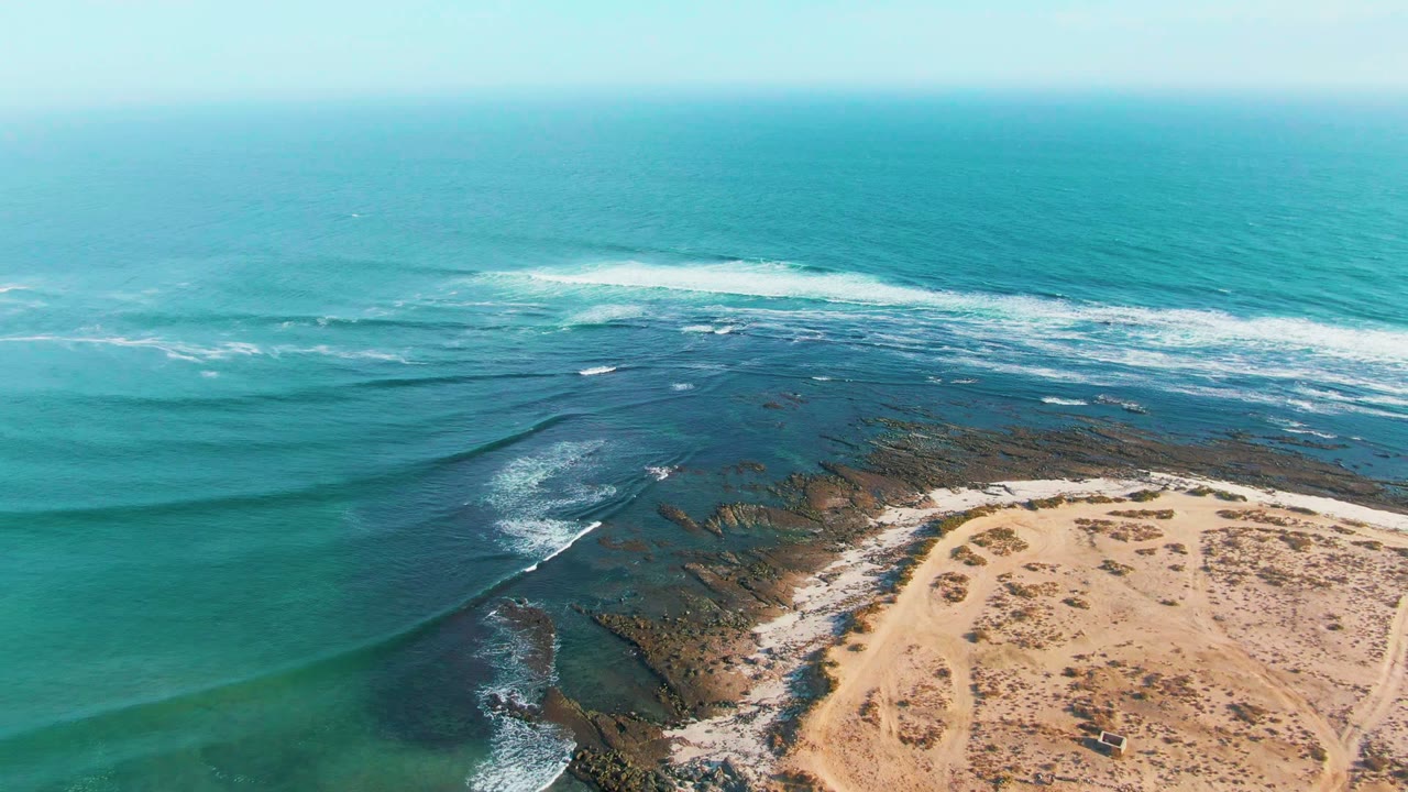 Drone view of white ocean foam breaking near dark coastal rocks and sand.