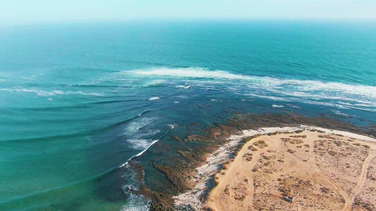 High aerial frame over La Bocana, Baja California Sur, with a broad turquoise water field, a breaking wave near the upper frame, a rocky edge, sandy ground, and no visible people.