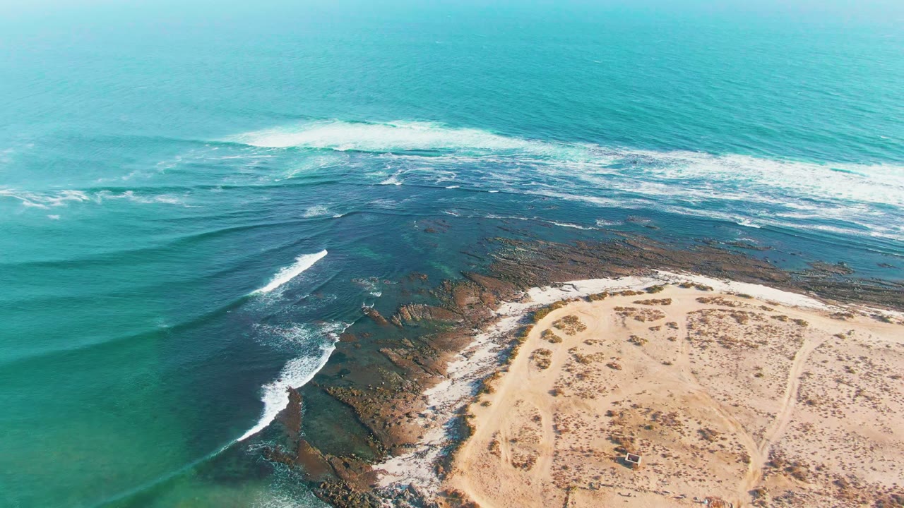 Opening aerial frame over La Bocana, Baja California Sur, with turquoise water, white surf on dark rocks, pale sand at the lower right, and no visible people.