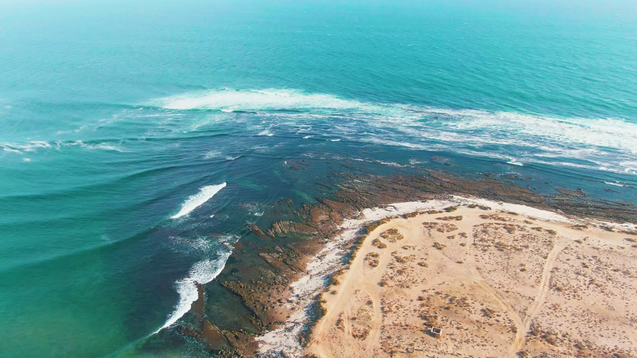 Drone view of waves rolling over dark rocks toward pale sand.