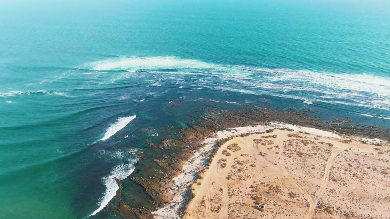 La Bocana coastline from above, with a longer white breaker crossing the dark rock shelf, open turquoise water beyond it, beach tracks at the lower right, and no visible people.
