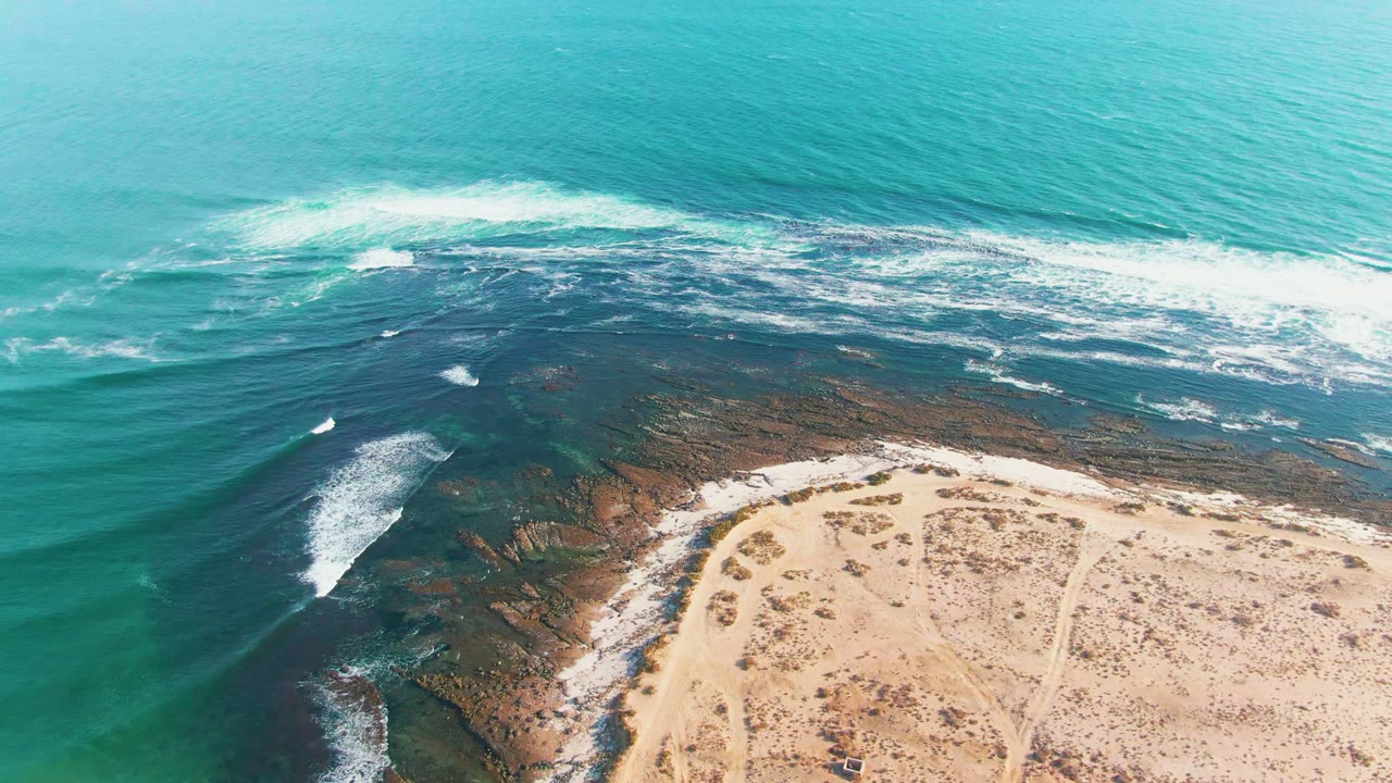Aerial frame over La Bocana, Baja California Sur, showing turquoise water, white surf on dark coastal rocks, tan sand at the bottom edge, and no visible people.