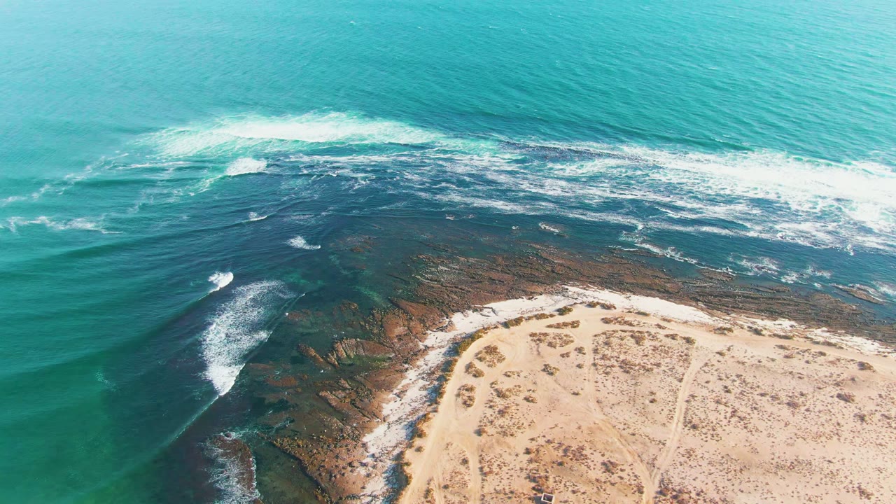 Aerial frame over La Bocana showing broken whitewater across the middle of the coast, darker reef under the water, pale beach in the lower frame, and no visible people.