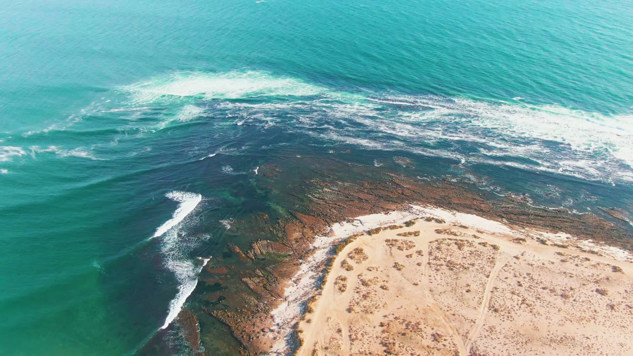 Aerial view of La Bocana coastline with turquoise water, white surf, dark reef, pale sand, and no visible people at the start of the clip.