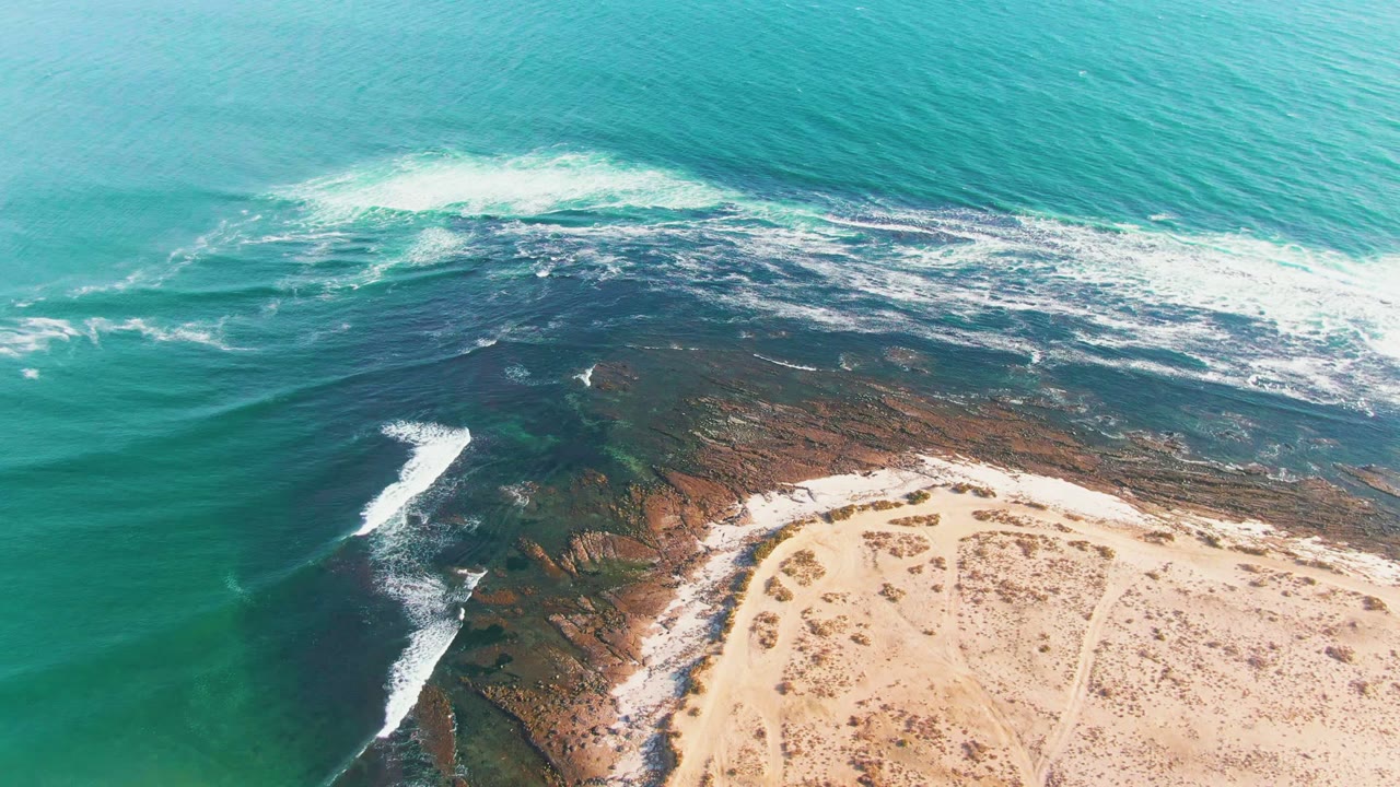 White foam spreads across dark shoreline rocks near a curved sandy beach.