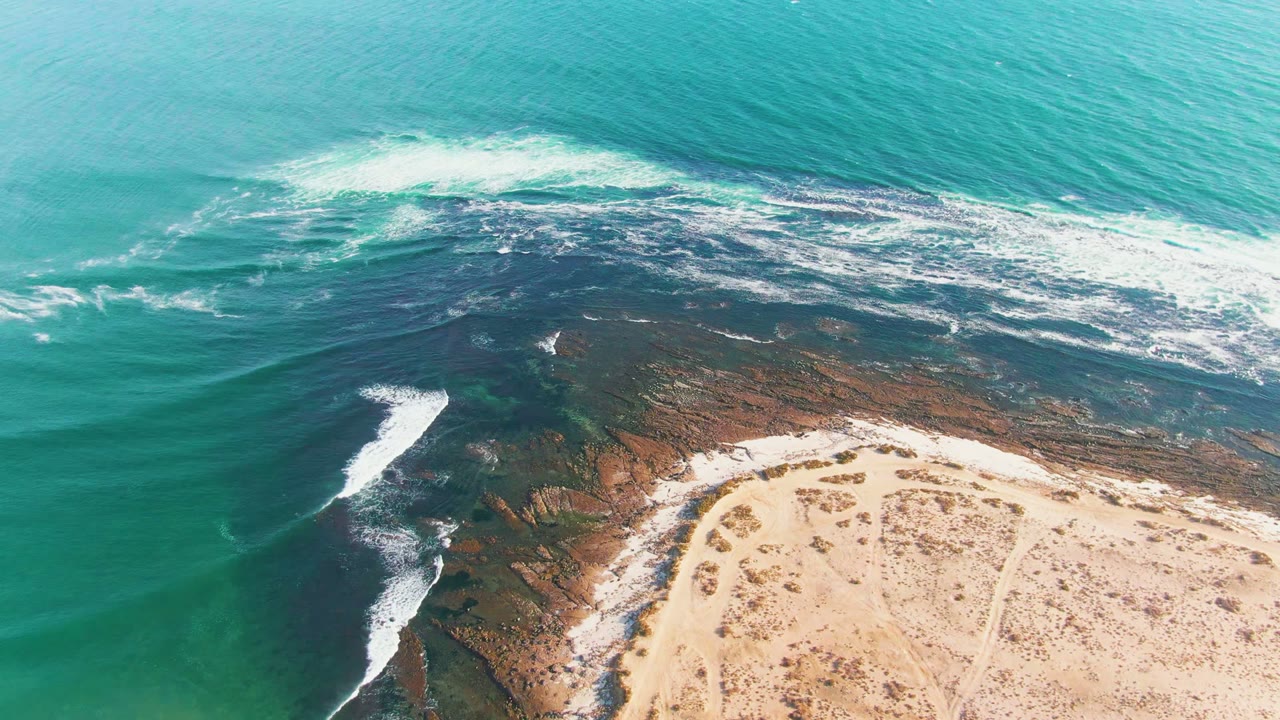 Aerial view of La Bocana surf wrapping around a rocky point beside light sand, with turquoise water above and no visible people.