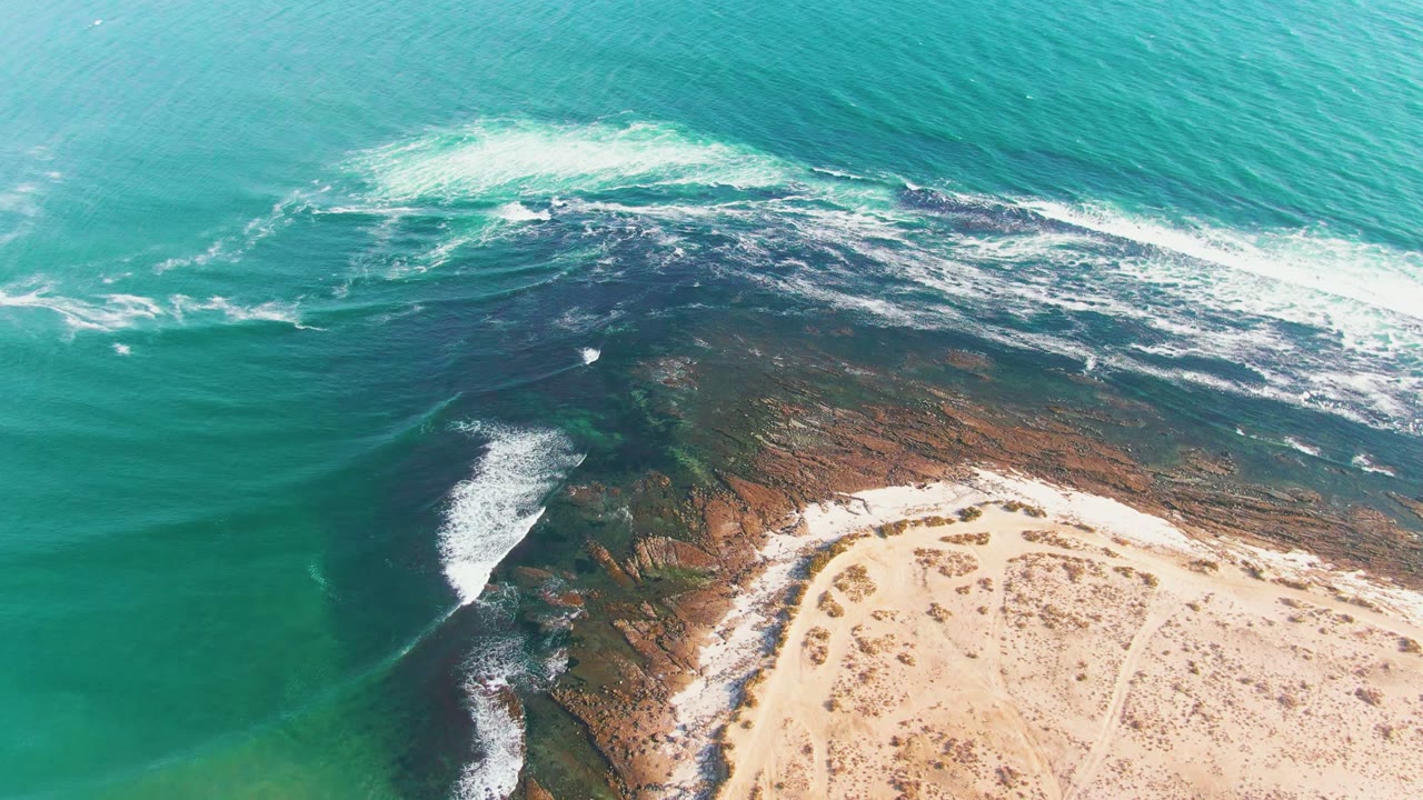 Aerial view at La Bocana, Baja California Sur, with turquoise water and white foam crossing a dark rocky shelf beside pale sand, no visible people.