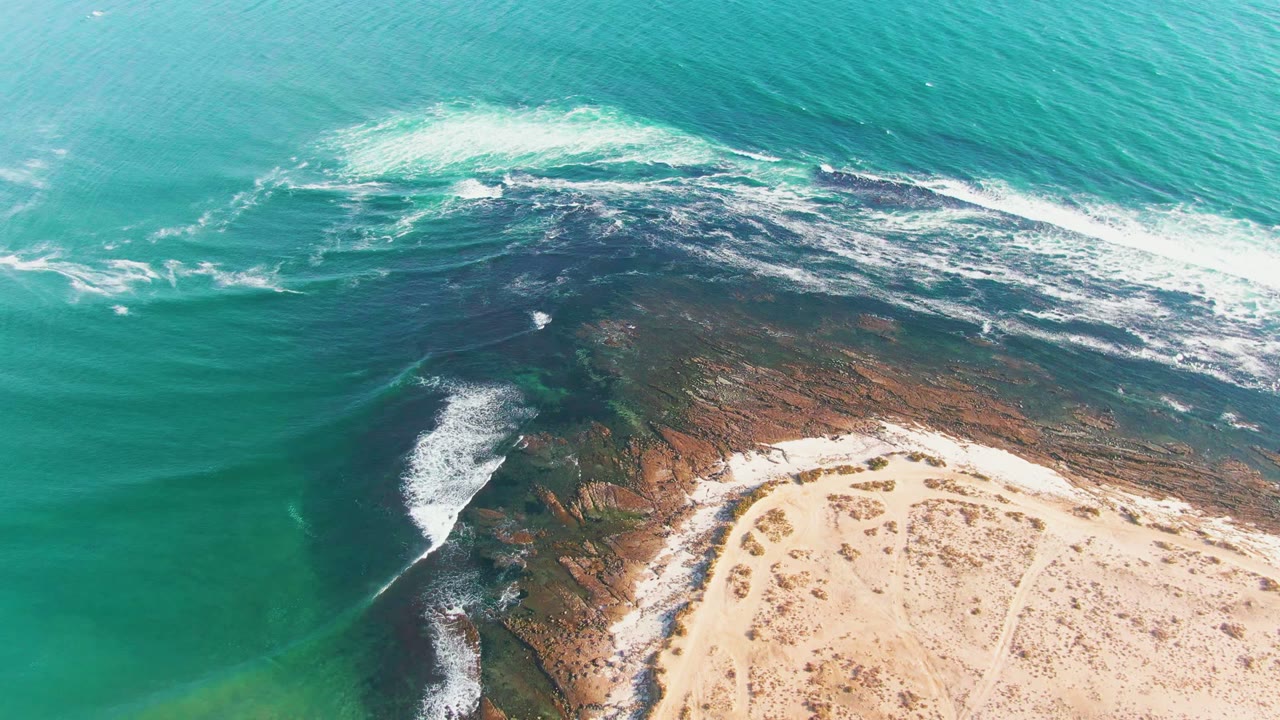 White surf spreads over dark coastal rocks near a pale beach.