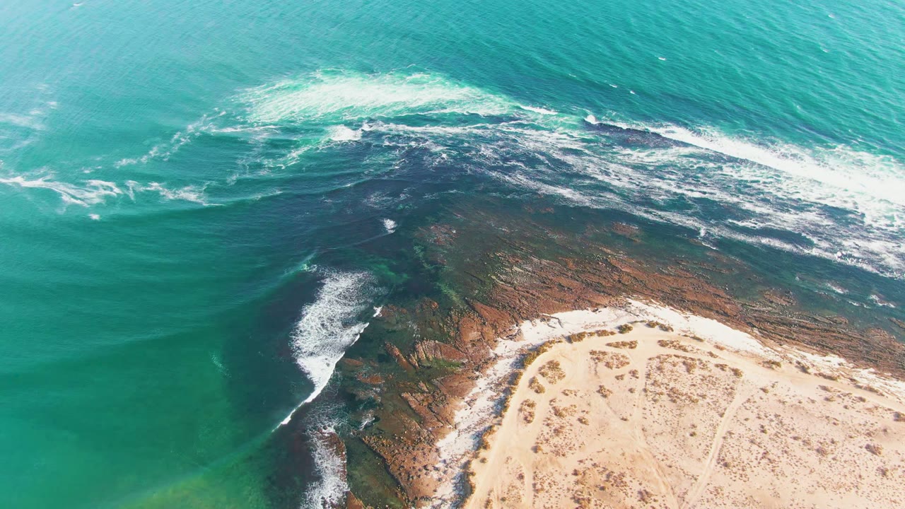 Aerial view over La Bocana coastline with green-blue water on the left, a foamy break over rocks, and the sandy shore curving through the lower right, no visible people.