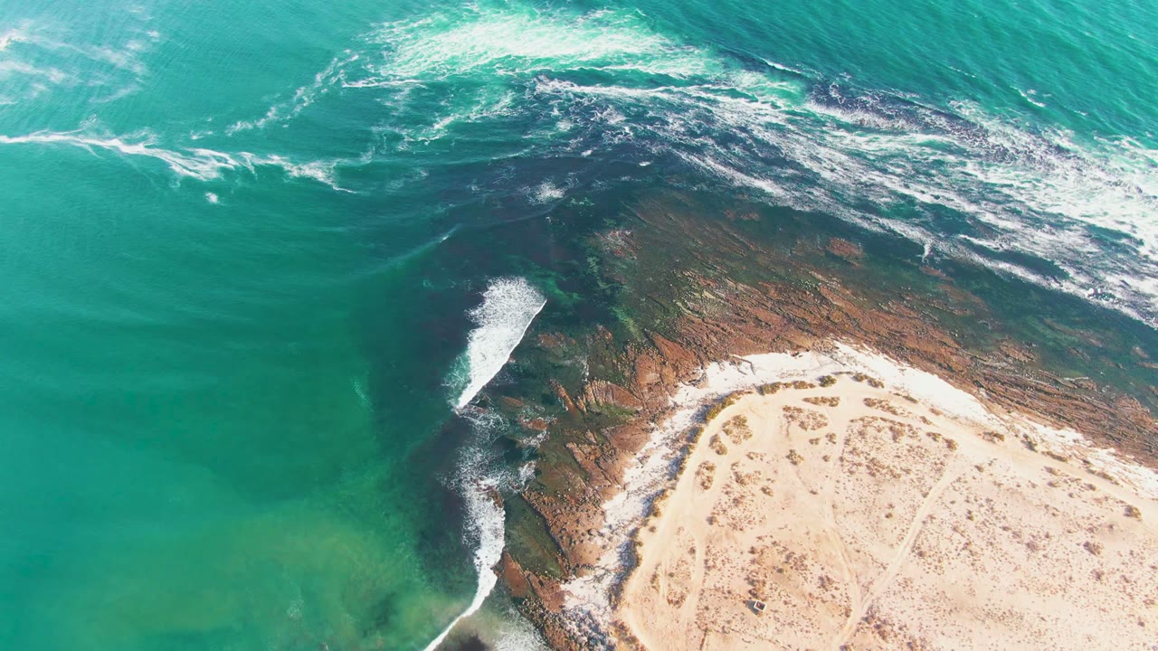 Aerial frame from La Bocana, Baja California Sur showing turquoise water, white foam, a dark rocky shelf, pale sand, and no visible people at the opening moment.