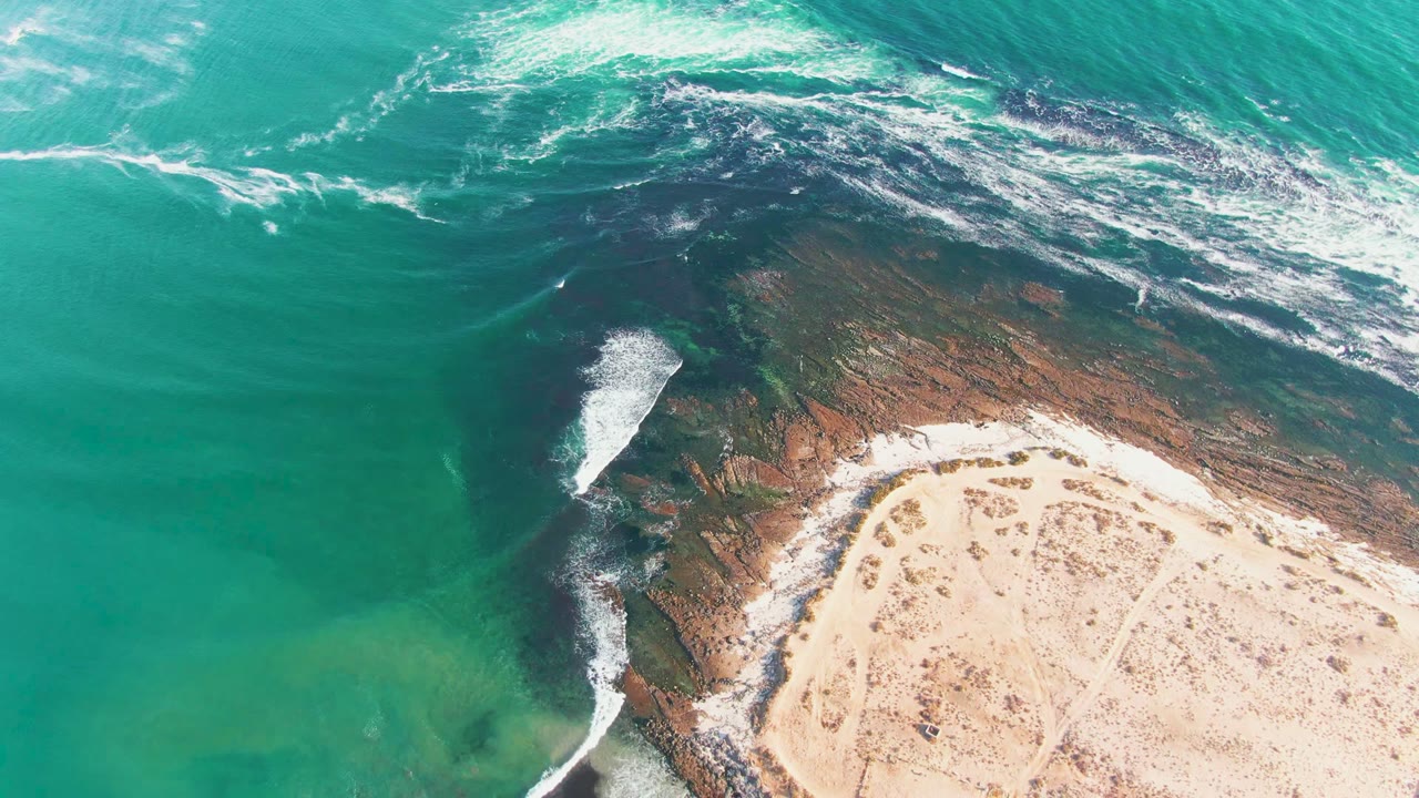 Waves wash across a rocky shoreline next to pale sand and green-blue water.