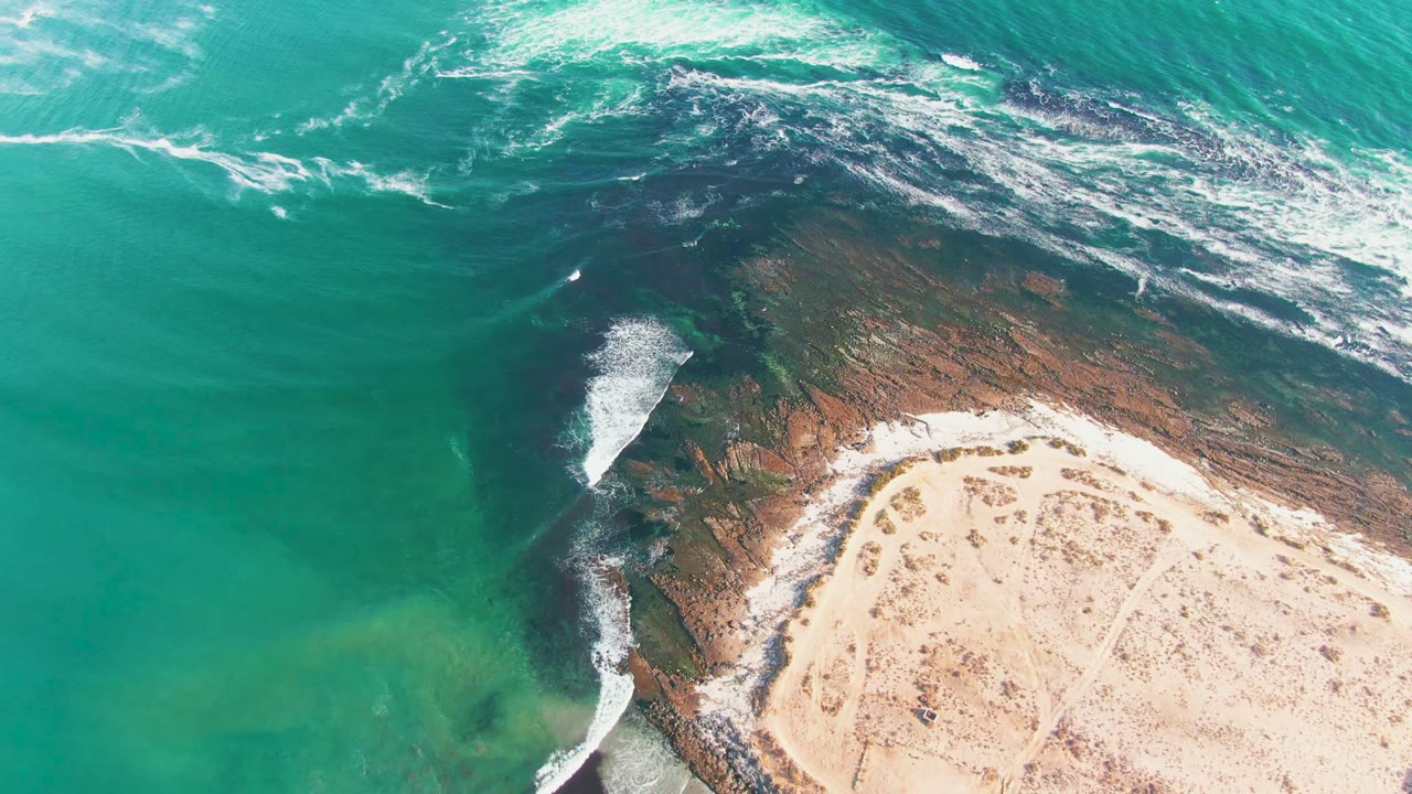 At 0:02 in La Bocana, long white foam lines sit near the top of the frame while the rocky point and pale beach remain visible below, with no visible people.