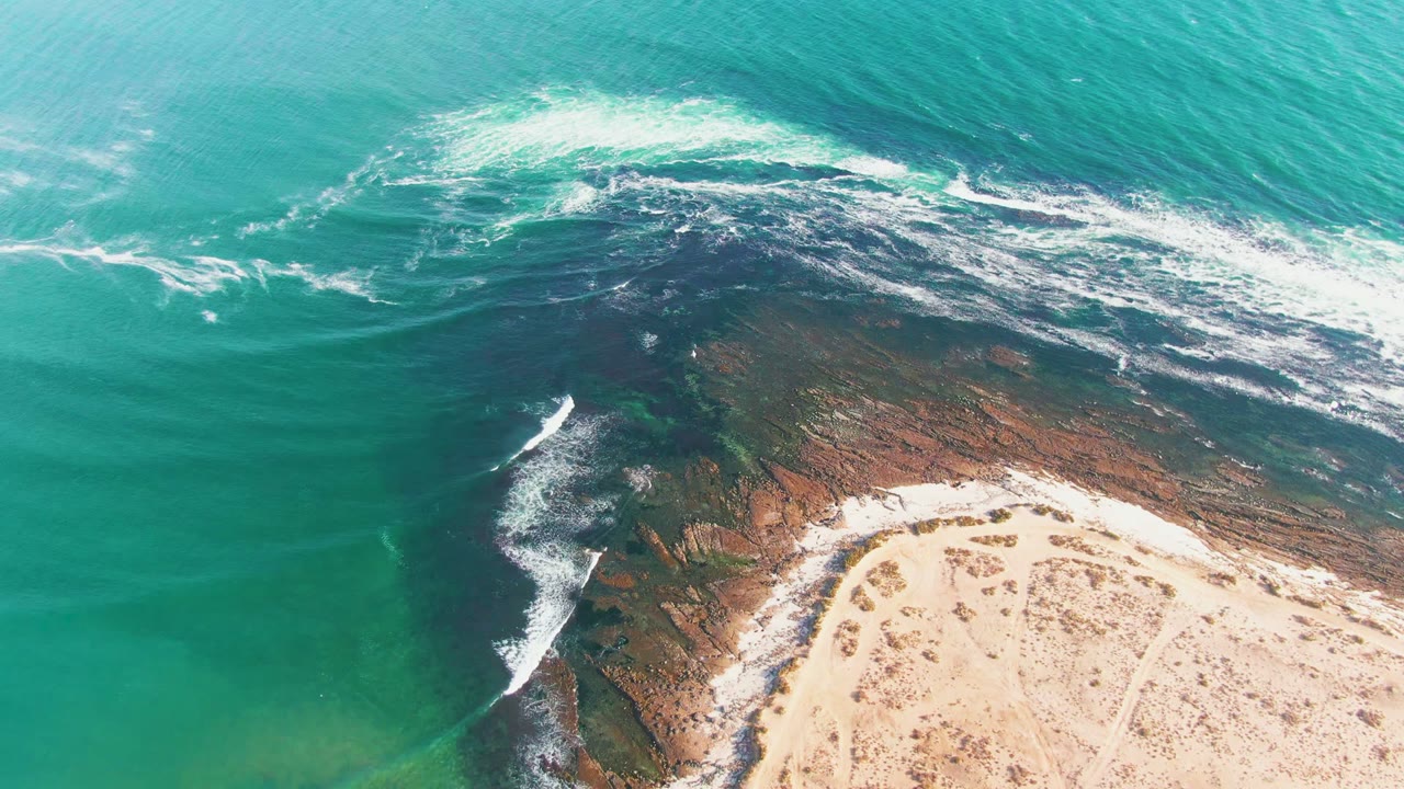 Aerial frame over La Bocana showing turquoise water, a dark rocky shelf, white surf, pale sand, and no visible people at the start of the clip.