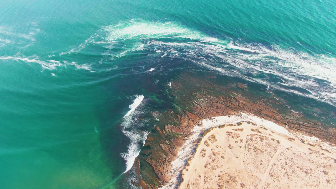 Aerial frame over La Bocana showing broader white foam across the upper rocky shelf, green-blue water at left, pale sand at bottom right, and no visible people.