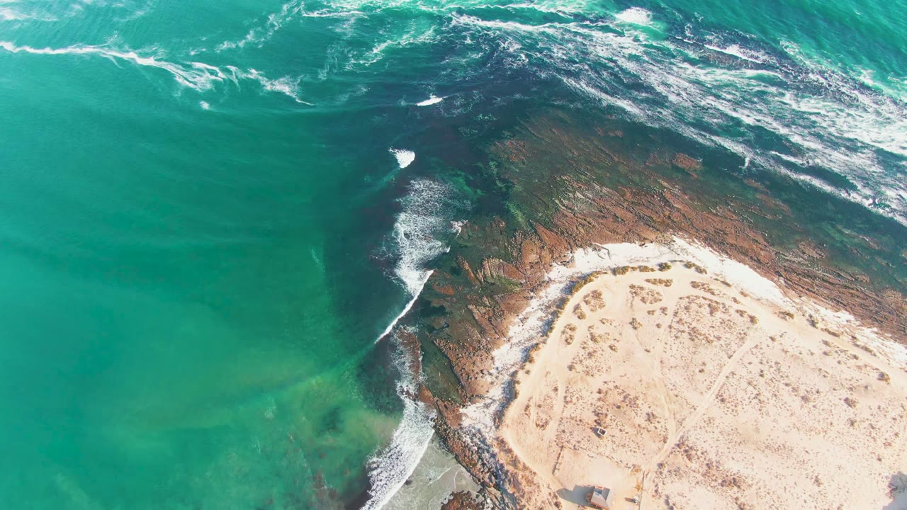 Aerial frame over La Bocana showing turquoise water, white surf on dark reef, pale sandy beach, and no visible people at the start of the clip.