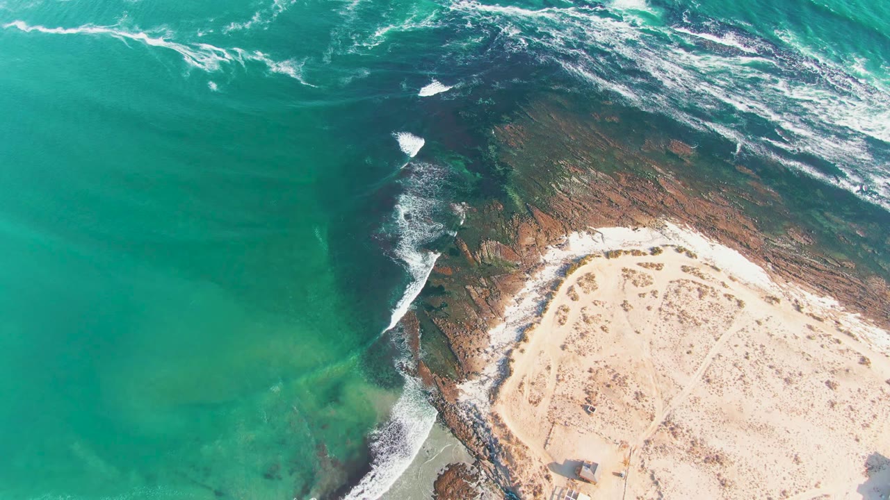 Aerial frame over La Bocana showing whitecaps streaking across the upper water, darker reef below, pale beach to the right, and no visible people.