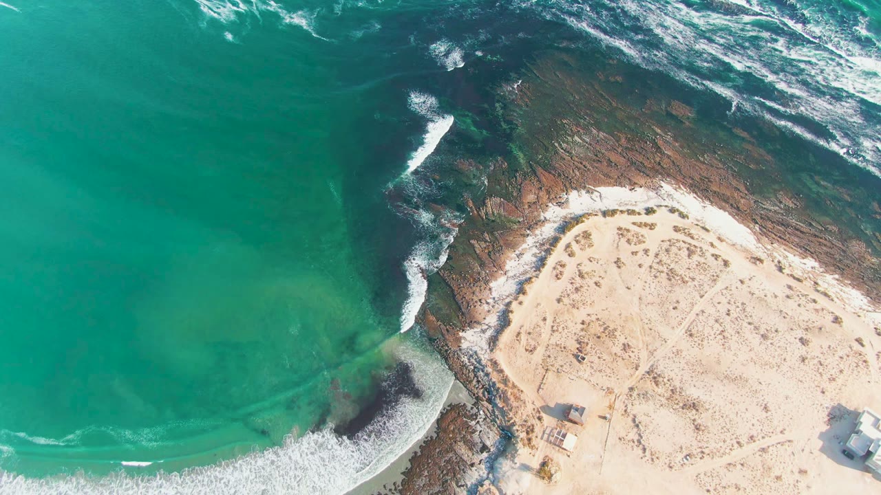 Aerial frame from La Bocana, Baja California Sur showing turquoise water, white surf, pale sand, dark shoreline rocks, small beach structures near the bottom edge, and no visible people.