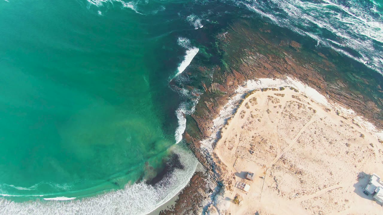 Overhead coastal view with waves curling around rocks beside a sandy beach.