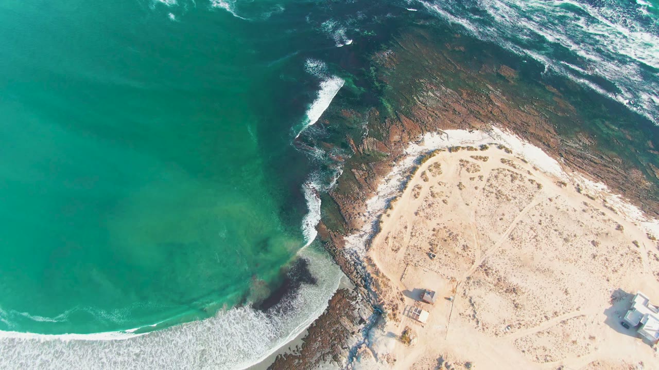 Overhead La Bocana coastline frame with broad turquoise water, whitecaps at the top, a dark rocky strip meeting pale beach sand, small structures at the lower right, and no visible people.