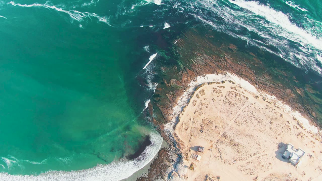 Aerial frame over La Bocana, Baja California Sur showing turquoise water meeting a dark rocky shelf and pale sand, with no visible people.