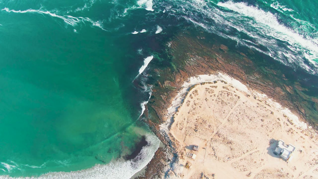Aerial frame over La Bocana with more white surf streaking across the upper rocks and a sandy point curving into the lower right, with no visible people.