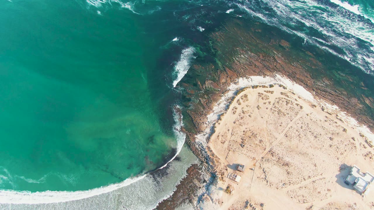 Overhead view in La Bocana, Baja California Sur, with turquoise water meeting a curved sandy beach and dark rocks, plus small structures on the sand and no visible people.