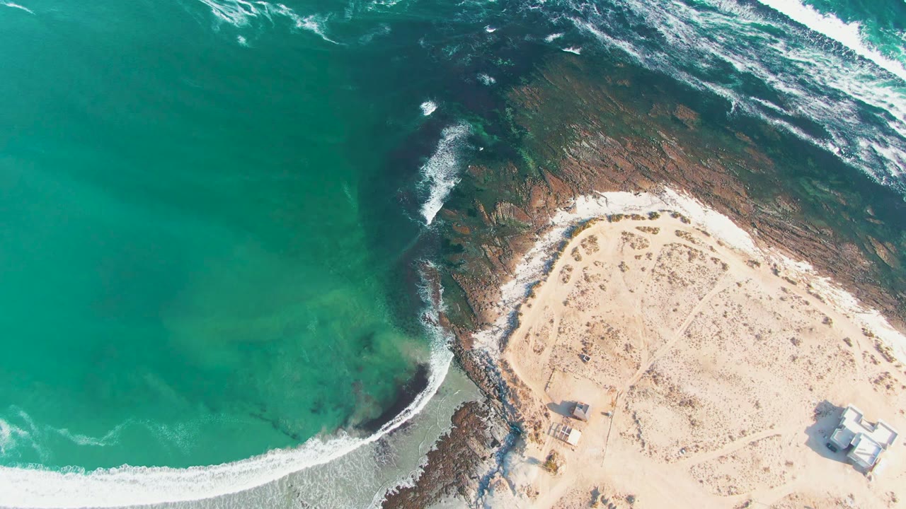 Top-down La Bocana coastline view with shallow turquoise water on the left, a dark rocky point through the center, bright sand on the right, and no visible people.