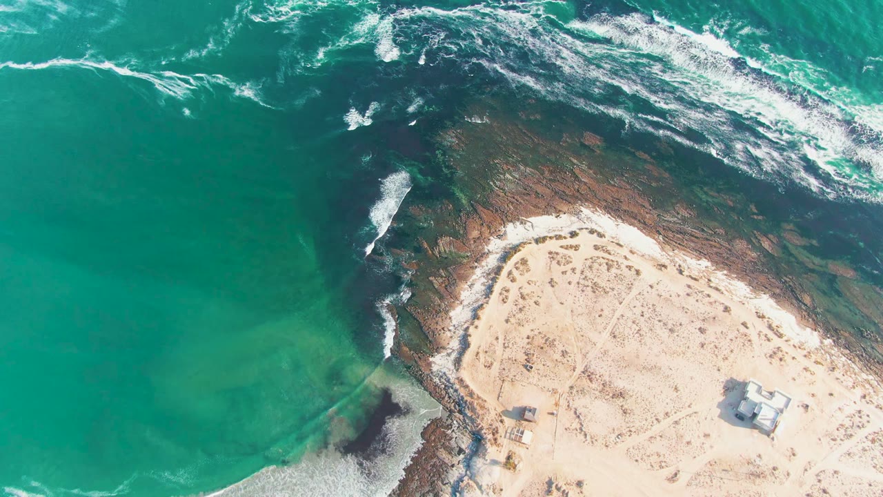 Opening overhead frame of La Bocana, Baja California Sur, with turquoise water at left, white surf crossing dark rock, pale sand at lower right, and no visible people.