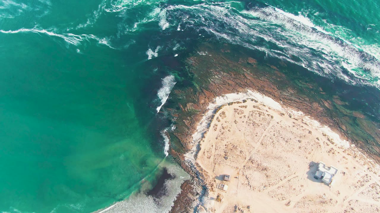 Aerial coastal frame with waves breaking over dark rocks beside pale sand.