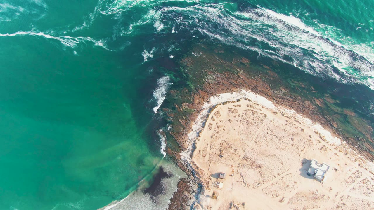 Aerial frame over La Bocana showing turquoise water wrapping the rocky point, a bright band of surf near the top, pale sandy ground, a small structure at lower right, and no visible people.
