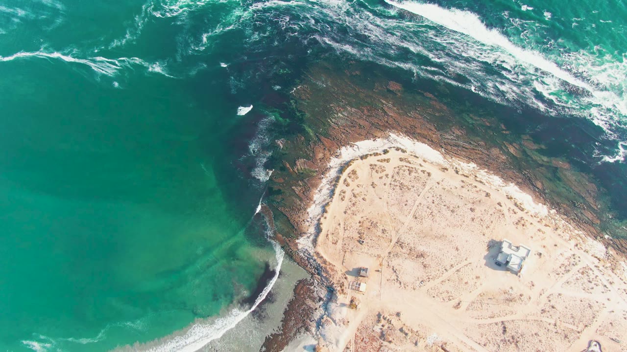 At 0:00, an overhead view of La Bocana shows turquoise water on the left, white surf crossing dark rocks, pale sand on the right, a small beach structure, and no visible people.