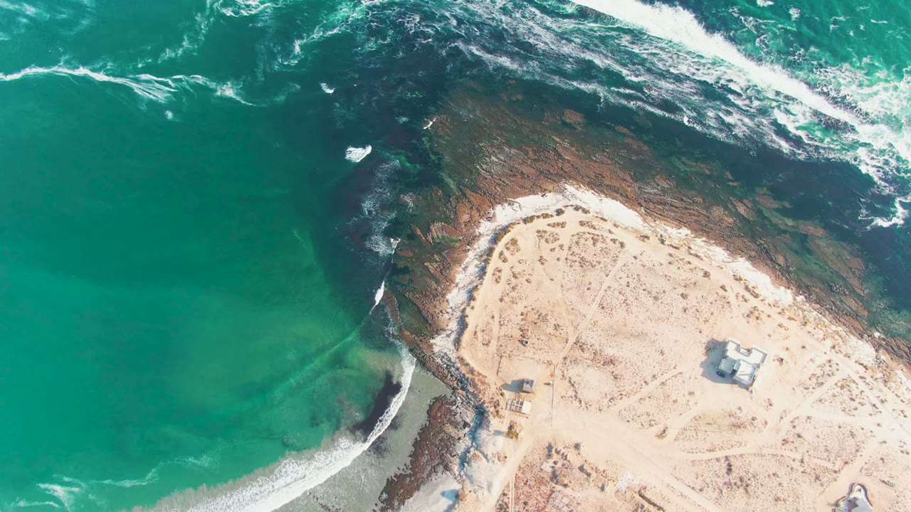 Aerial shoreline with white foam sliding around rocks beside pale sand.
