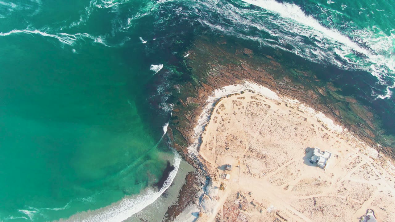 At 0:02, the overhead La Bocana view shows fresh whitewater near the upper surf band, exposed dark rock below it, pale sand at lower right, and no visible people.