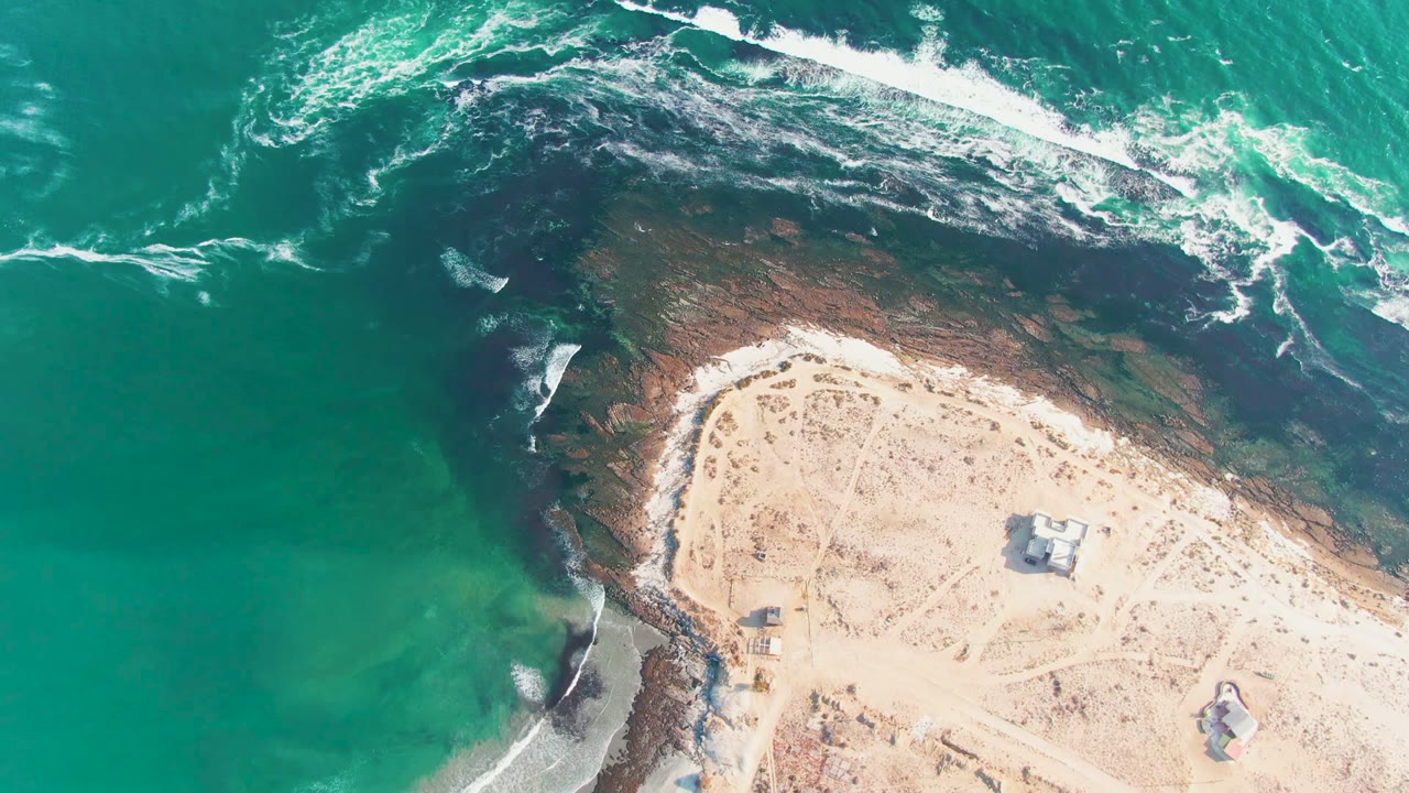 Aerial frame over La Bocana, Baja California Sur, Mexico, with turquoise water at left, white surf across dark rocks, pale sand at lower right, small structures, and no visible people.