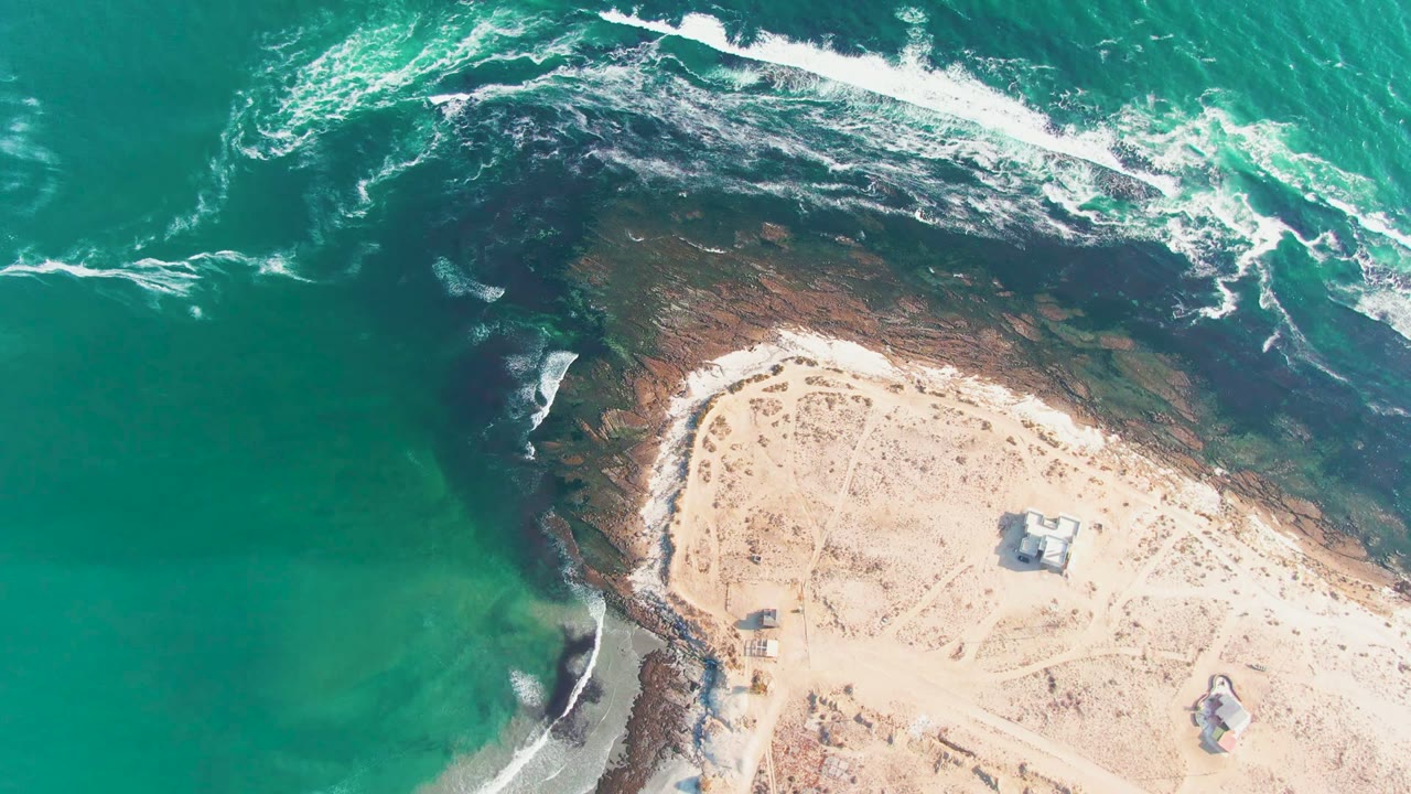 White wave foam spreads across a dark rocky shoreline next to pale sand.