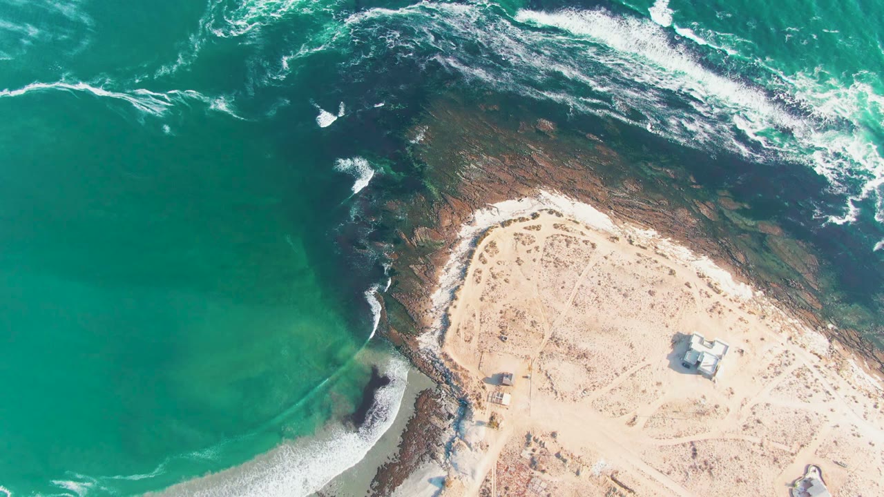 Aerial frame over La Bocana, Baja California Sur showing turquoise ocean, white foam, dark reef, pale sand, a small shore structure, and no visible people at the start of the clip.