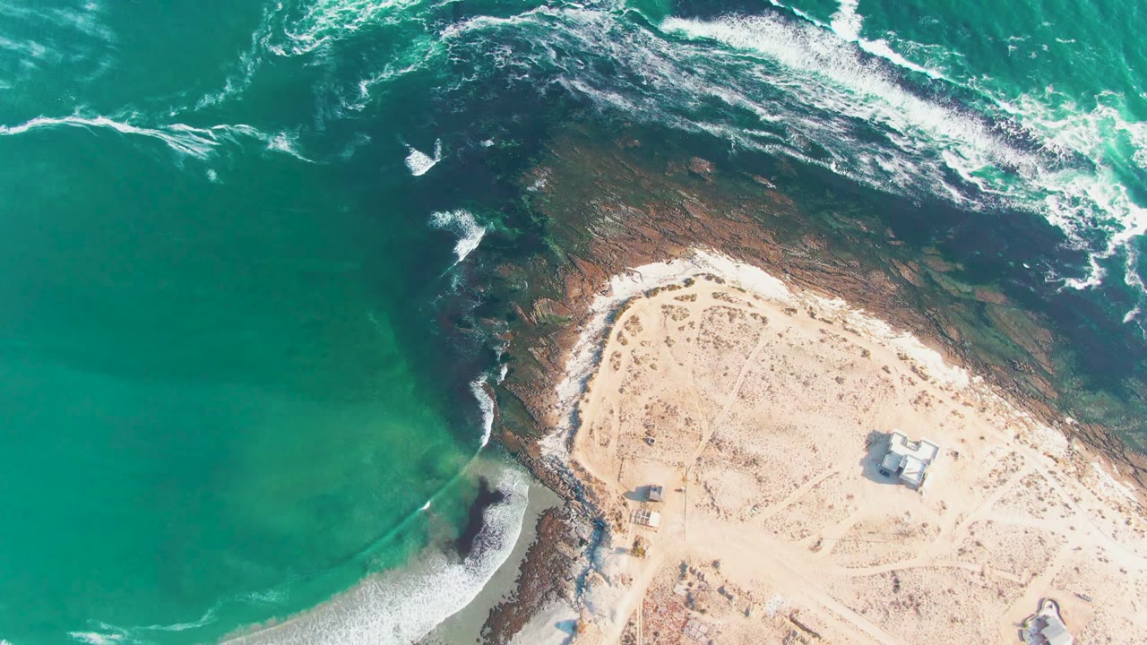 Foamy waves curl along a dark reef beside a pale sandy beach.
